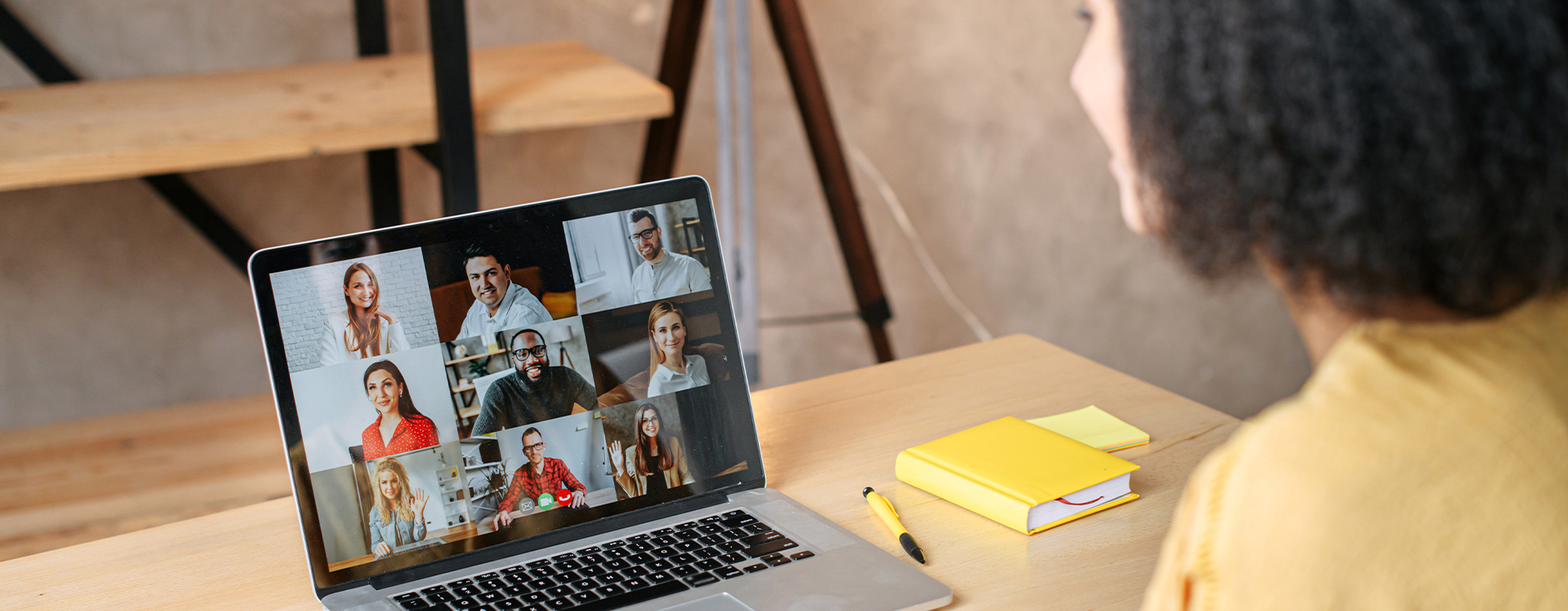 Woman in a yellow shirt sitting at a table with a laptop that has 7 colleagues on a video call.