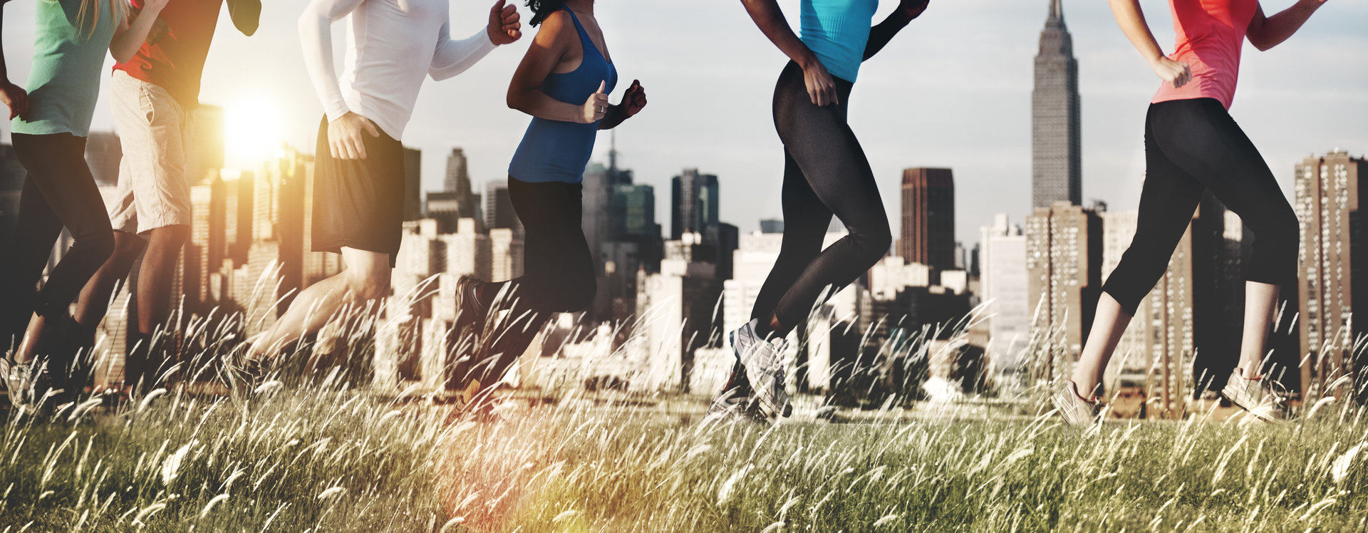 Line of people in workout clothes running in a grassy field with a city skyline behind them.