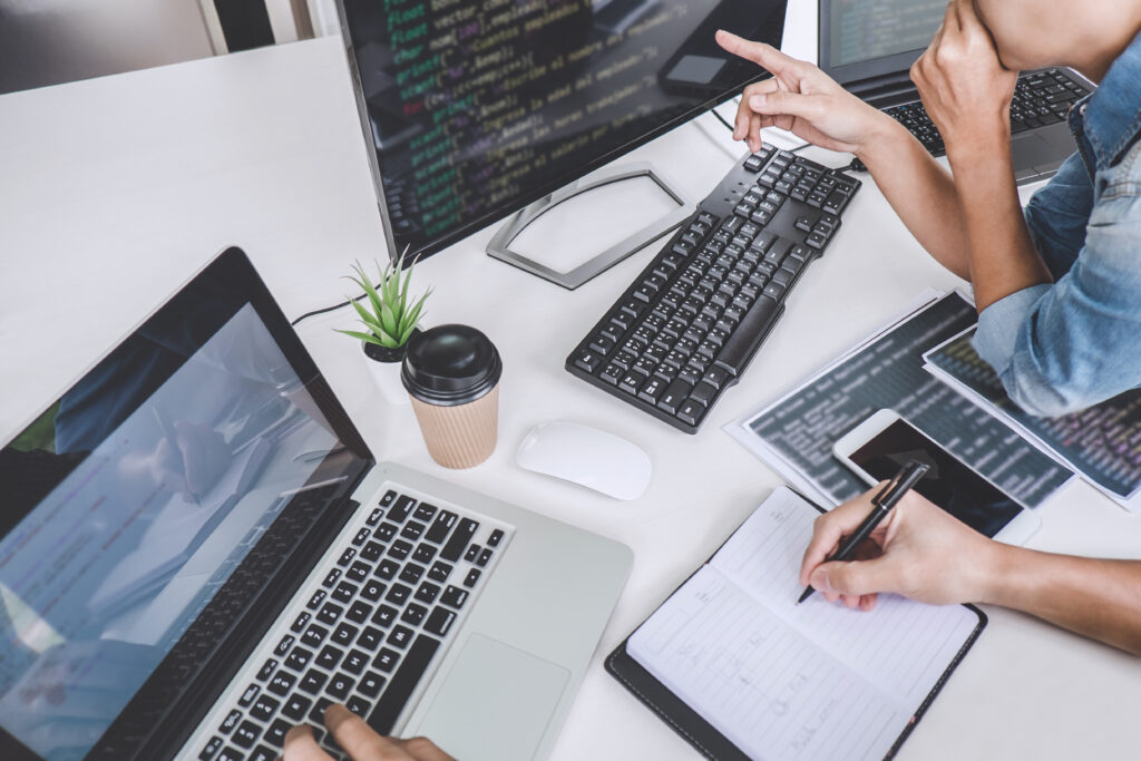 Person writing in journal next to woman pointing at monitor with coding on it.