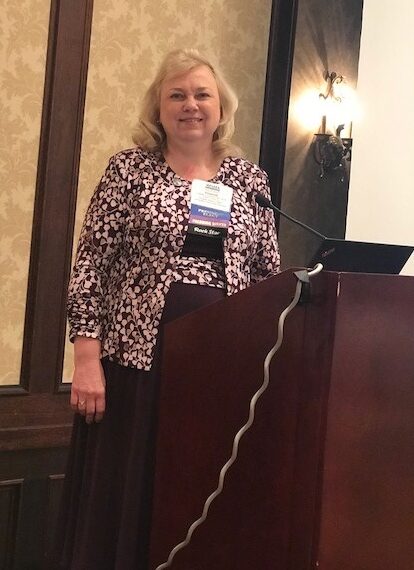 UW HIMT program director and capstone instructor Betty Rockendorf standing and smiling in front of a podium at a conference. 