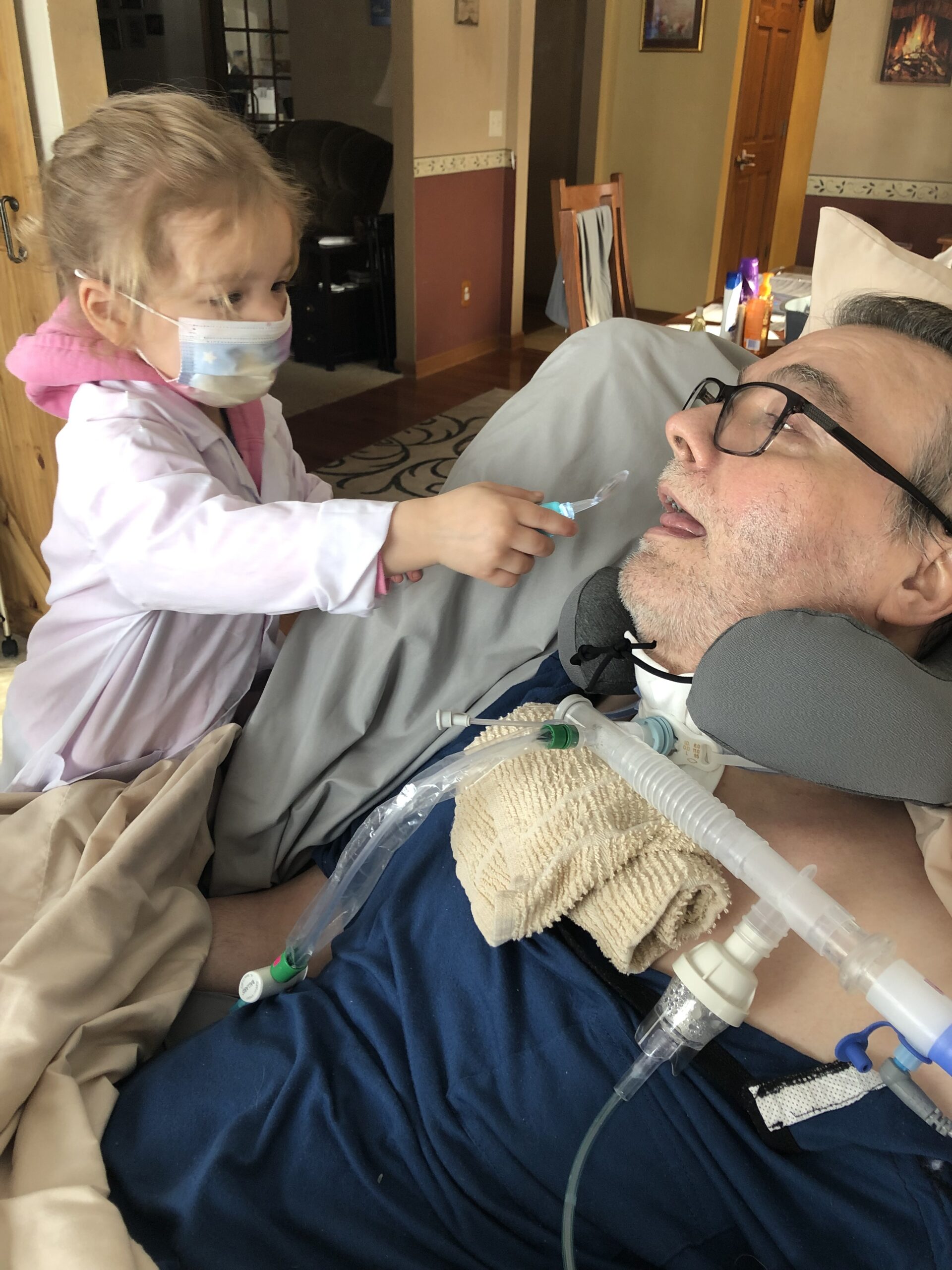 Britni's daughter wears a cloth mask as she readies a medical tube next to Britni's father.