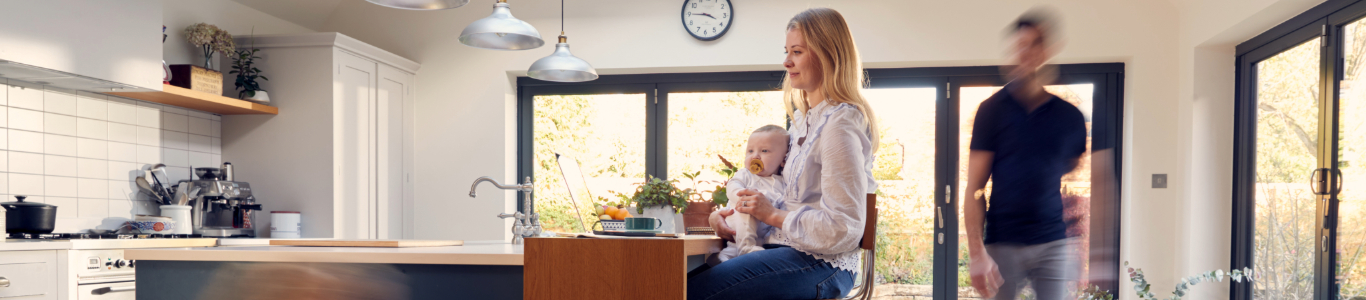 busy mom studying on her laptop holding a baby in her lap while her child and husband walk through the kitchen