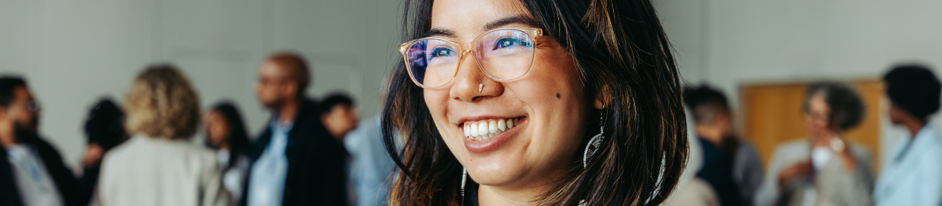 Smiling businesswoman wearing glasses at a biotechnology networking conference with colleagues in the background.