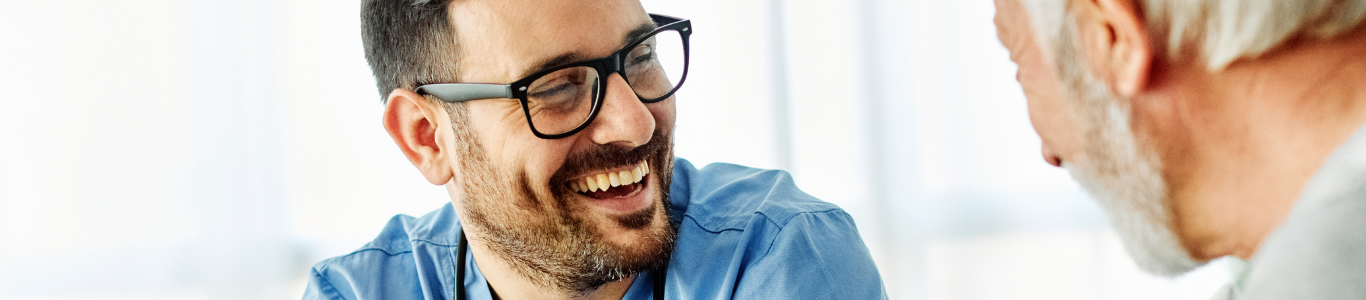 A photo of a male nurse with glasses smiles as he talks to an elderly man