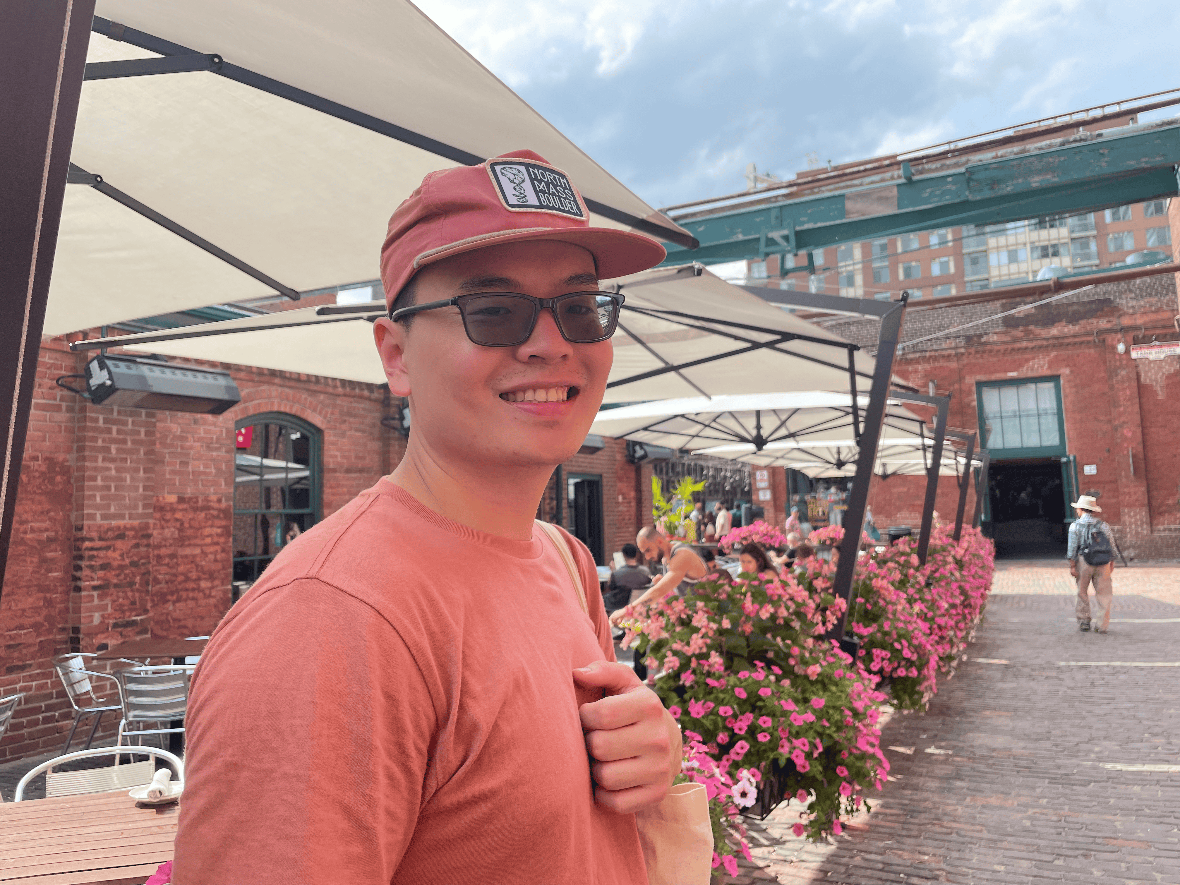 Applied Biotechnology graduate Jason Muljadi in a red shirt and cap standing near outdoor cafe seating with pink flowers and brick buildings in the background.