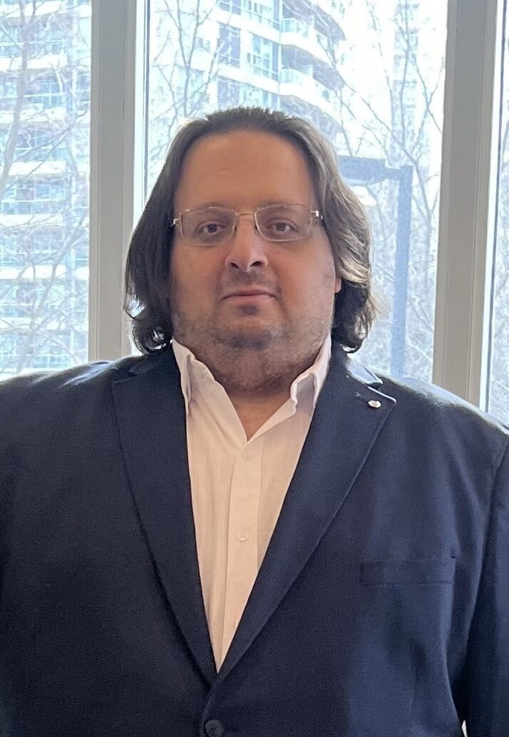 A picture of Abdallah Al Alfy, a graduate of the online Applied Biotechnology master’s program. Alfy wears a suit as he stands in front of a window overlooking a high rise building. 