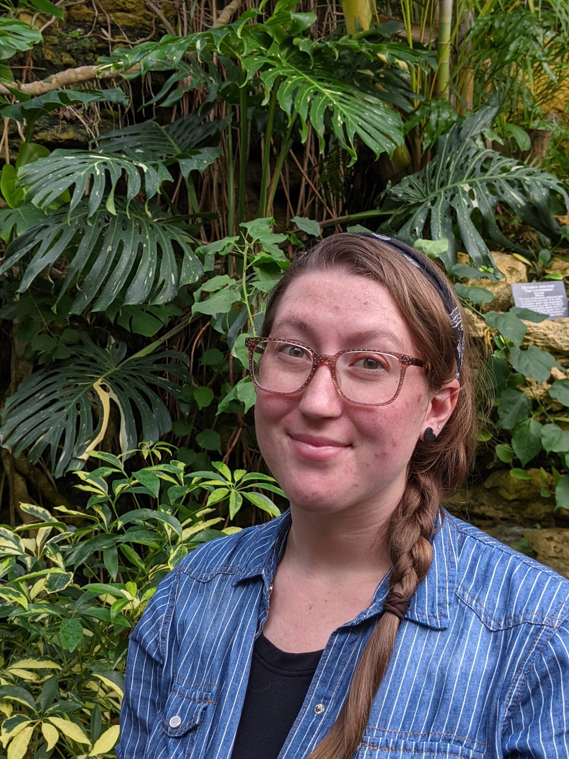Hazel with glasses and braided hair smiling in front of lush tropical plants with large green leaves.