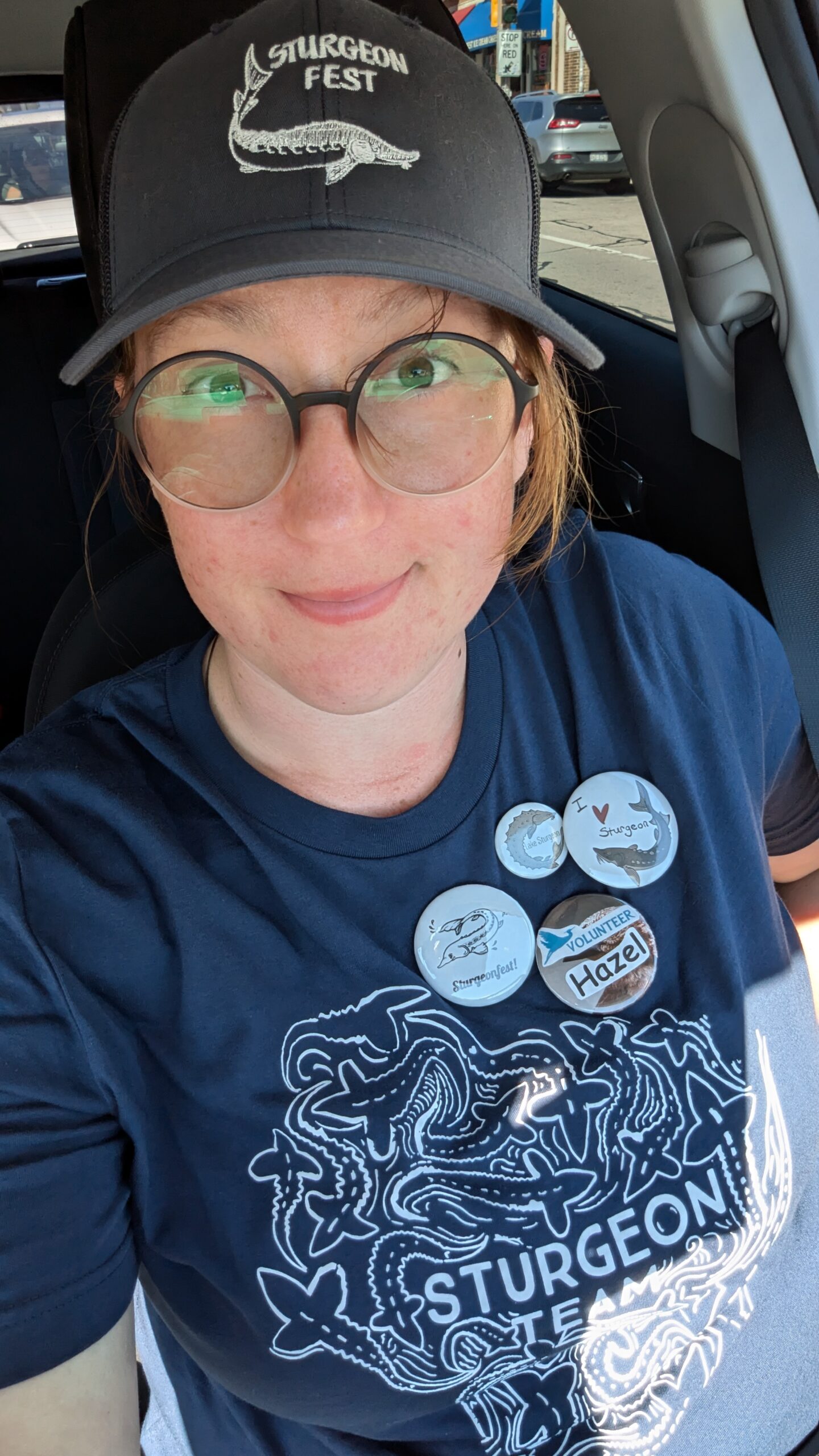 Hazel wearing a black Sturgeon Fest cap and matching navy blue Sturgeon Fest T-shirt with multiple event pins attached.