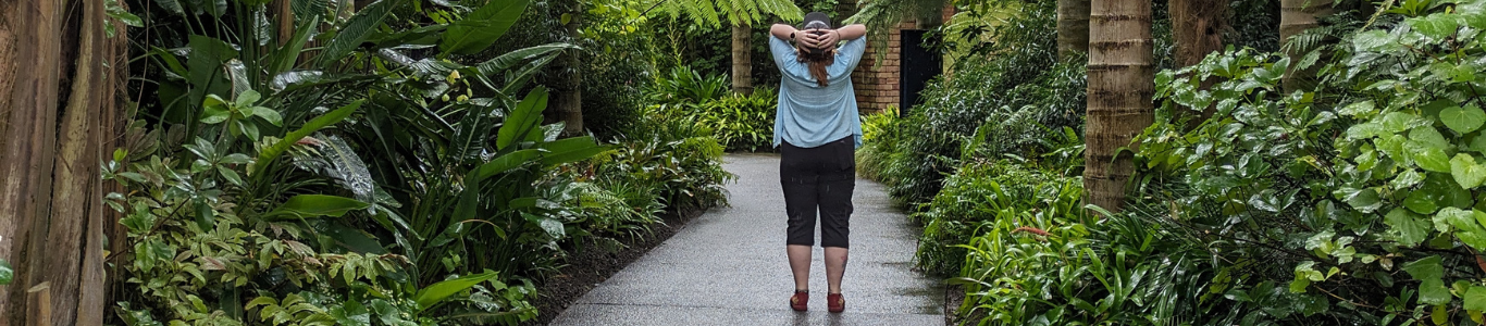 Sustainable Management student Hazel Ratka standing on a wet pathway surrounded by lush tropical plants and tall palm trees in a serene garden setting.