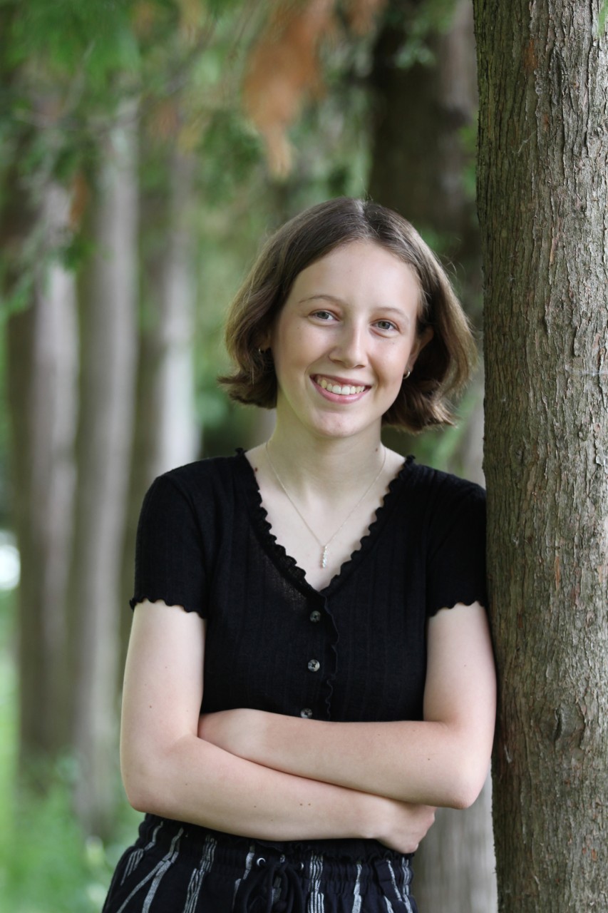 Karly smiling and leaning against a tree in a forested area wearing a black short-sleeve top.