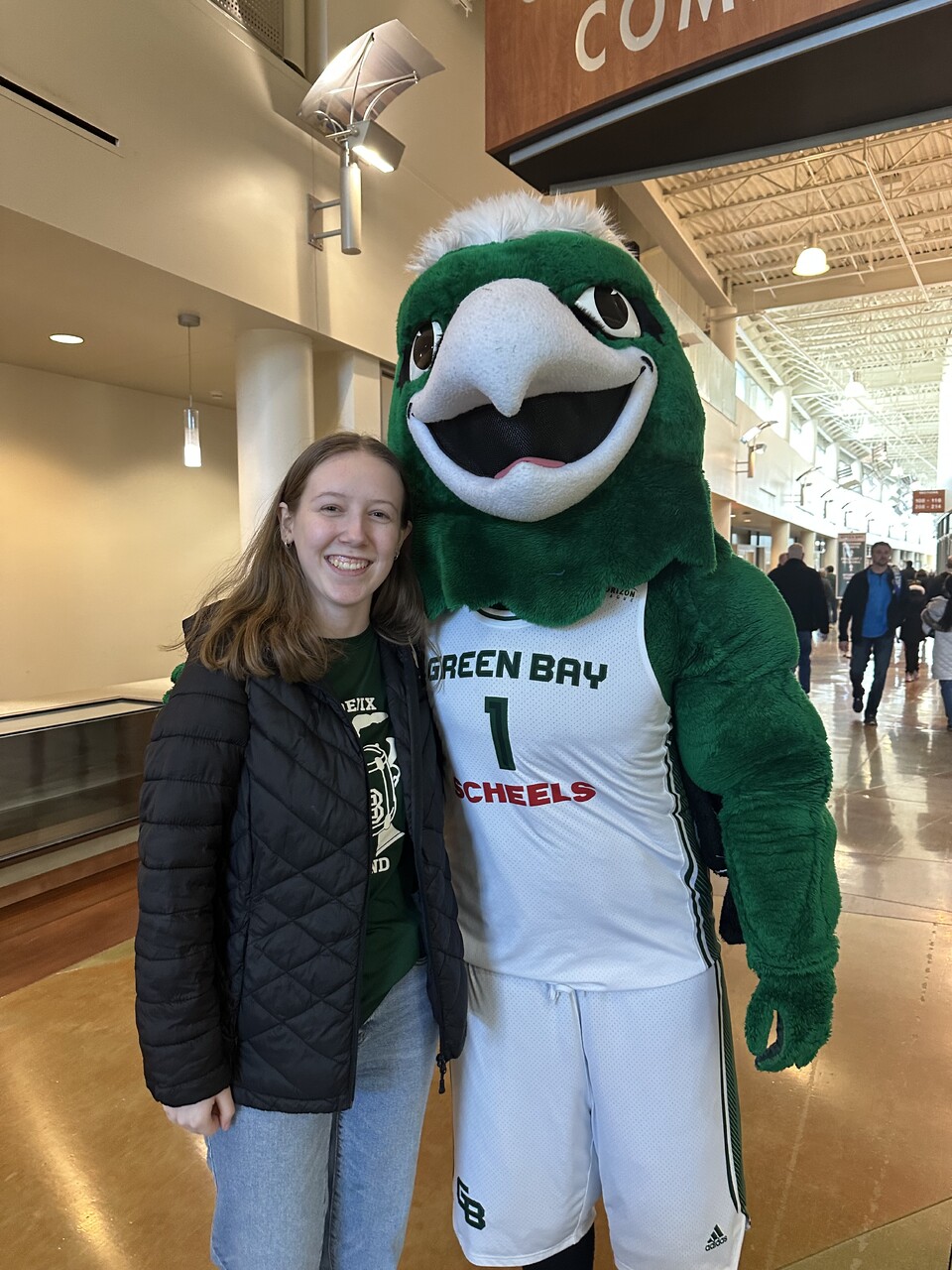 Karly smiling next to the UW-Green Bay basketball team mascot in a sports arena concourse.